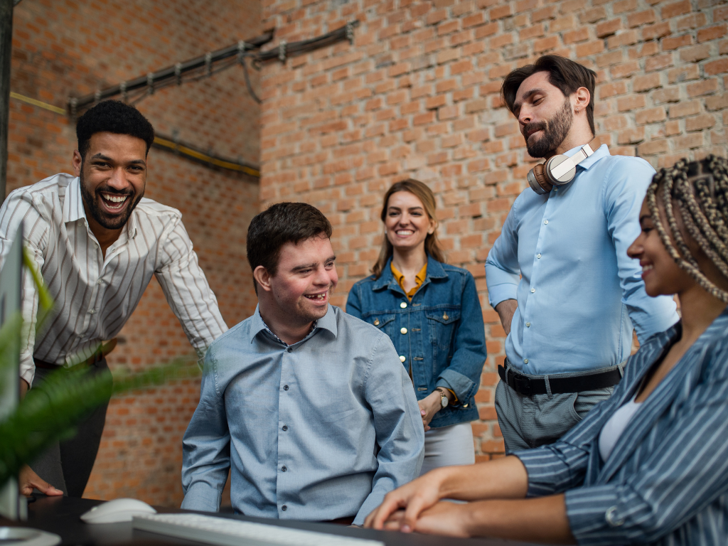A diverse group of five colleagues gathers around a computer, smiling and engaging in a lively discussion in an office with exposed brick walls. The group includes three men and two women, all appearing relaxed and collaborative, suggesting a positive and inclusive work environment.