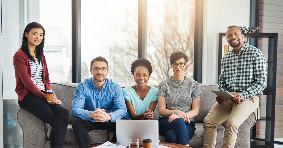 A diverse team of five professionals sitting together in a modern office space, smiling at the camera. They are gathered around a laptop with documents and coffee cups on the table, suggesting a collaborative work environment. This represents the teamwork and efficiency supported by AxiomEHR in behavioral health organizations.