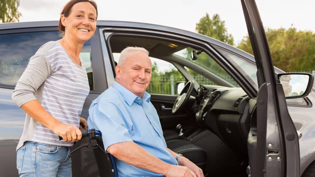 A woman is helping a man in a wheelchair into a van as a part of whole-person care.