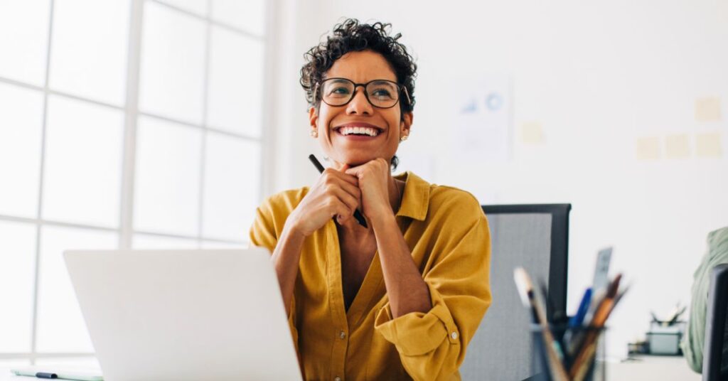 woman at her desk with a laptop smiling with hands clasped under her face