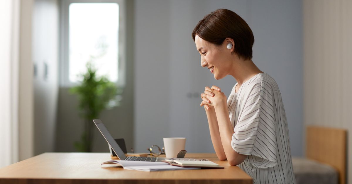 A woman providing behavioral health telehealth sits at a table smiling at a laptop.