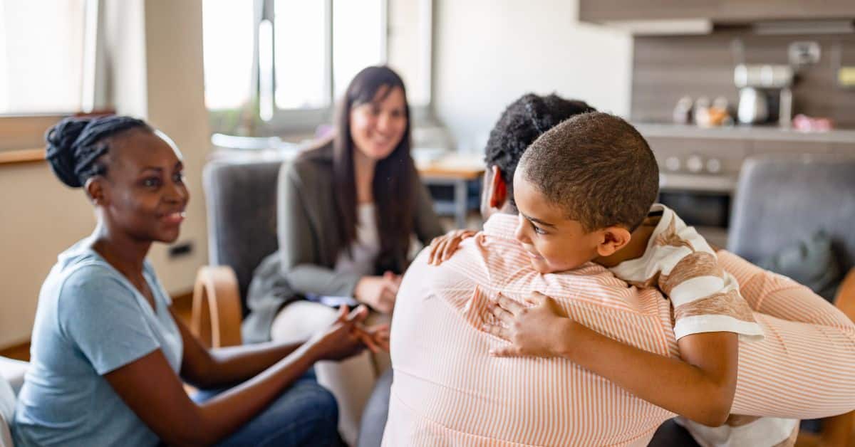 A child hugs a caregiver during a family support meeting with social workers in a home setting, reflecting discussions and updates related to medicaid news.