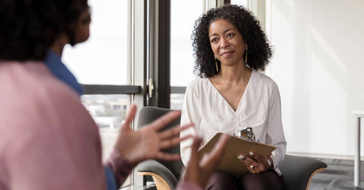 Counselor holding a clipboard listens attentively to a client speaking during a session, while gathering Medicaid work requirements documentation.